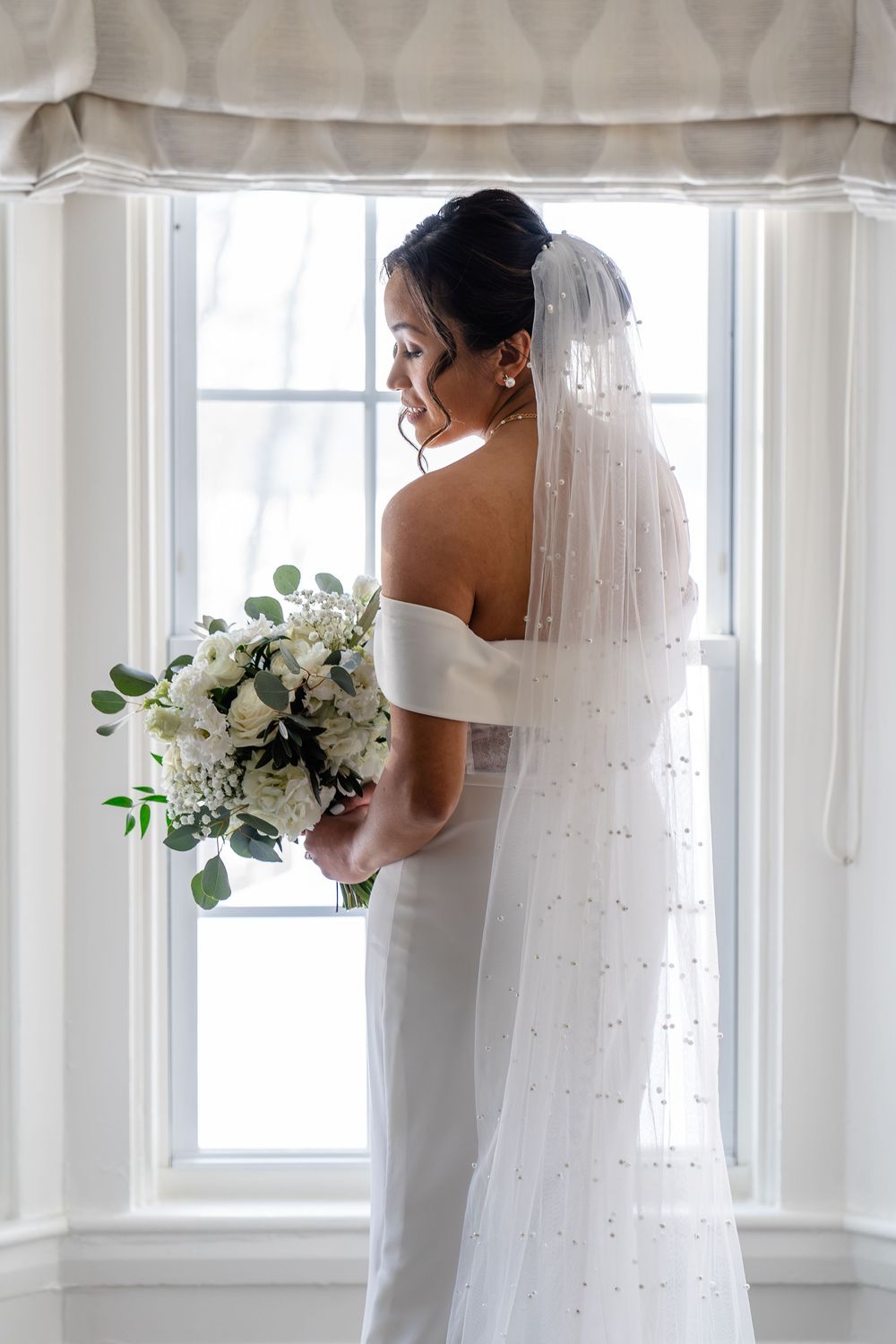 Bridal portrait by window with pearl veil and white floral bouquet at The Saphire Estate