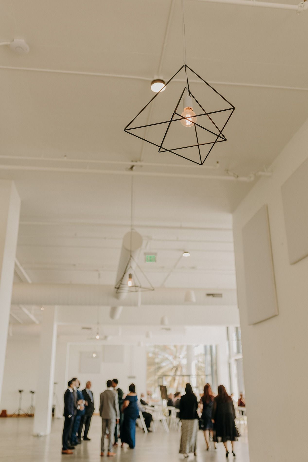 Geometric black wire pendant light hanging in a bright modern gallery space with blurred guests mingling below at The Lane.