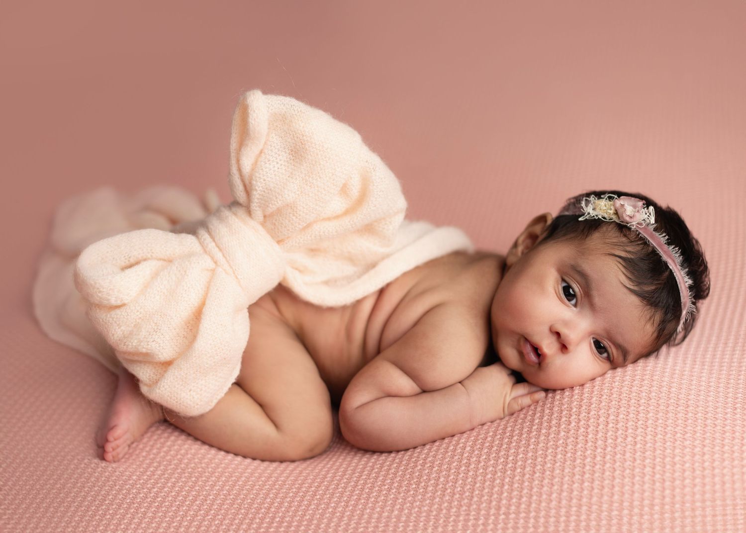 Newborn baby girl lying on pink blanket wearing a floral headband and large cream bow, in a professional photo.
