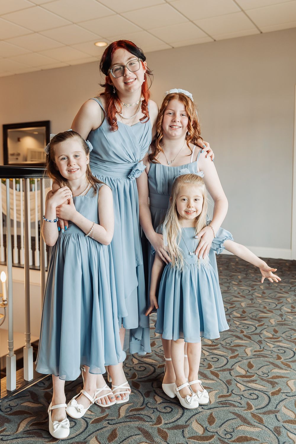 Woman and three girls in matching dusty blue dresses posing together on a staircase landing at a formal event.