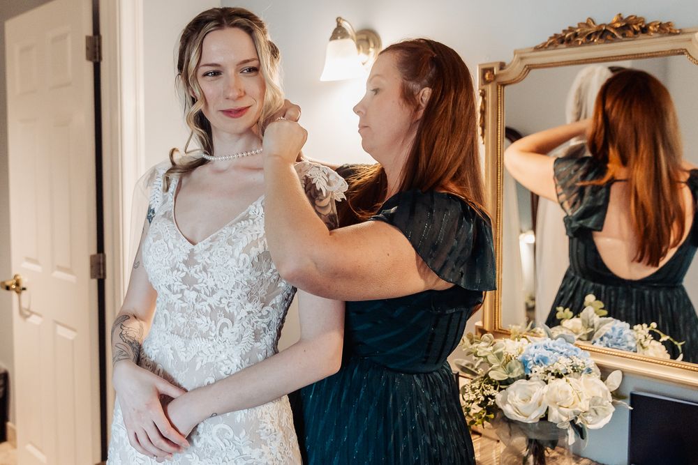 A bride in a lace wedding gown gets her dress fastened by a bridesmaid in a dark dress, reflected in a gold mirror.
