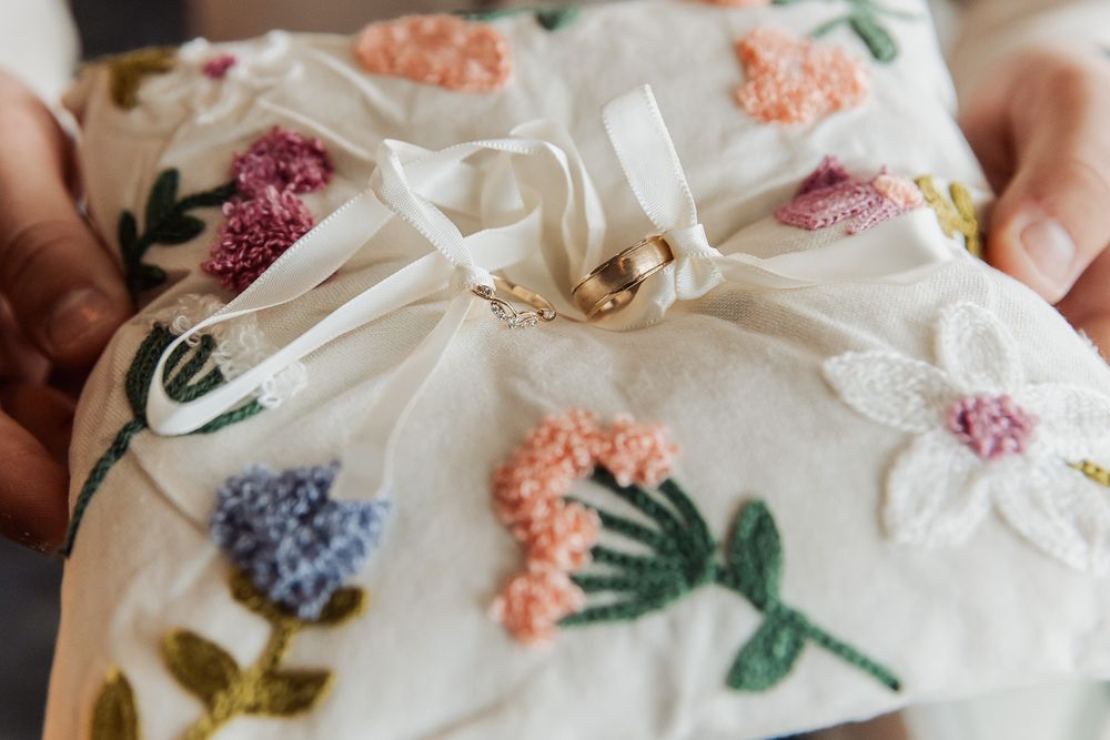 Two gold wedding rings resting on an embroidered floral ring bearer pillow held by a person.