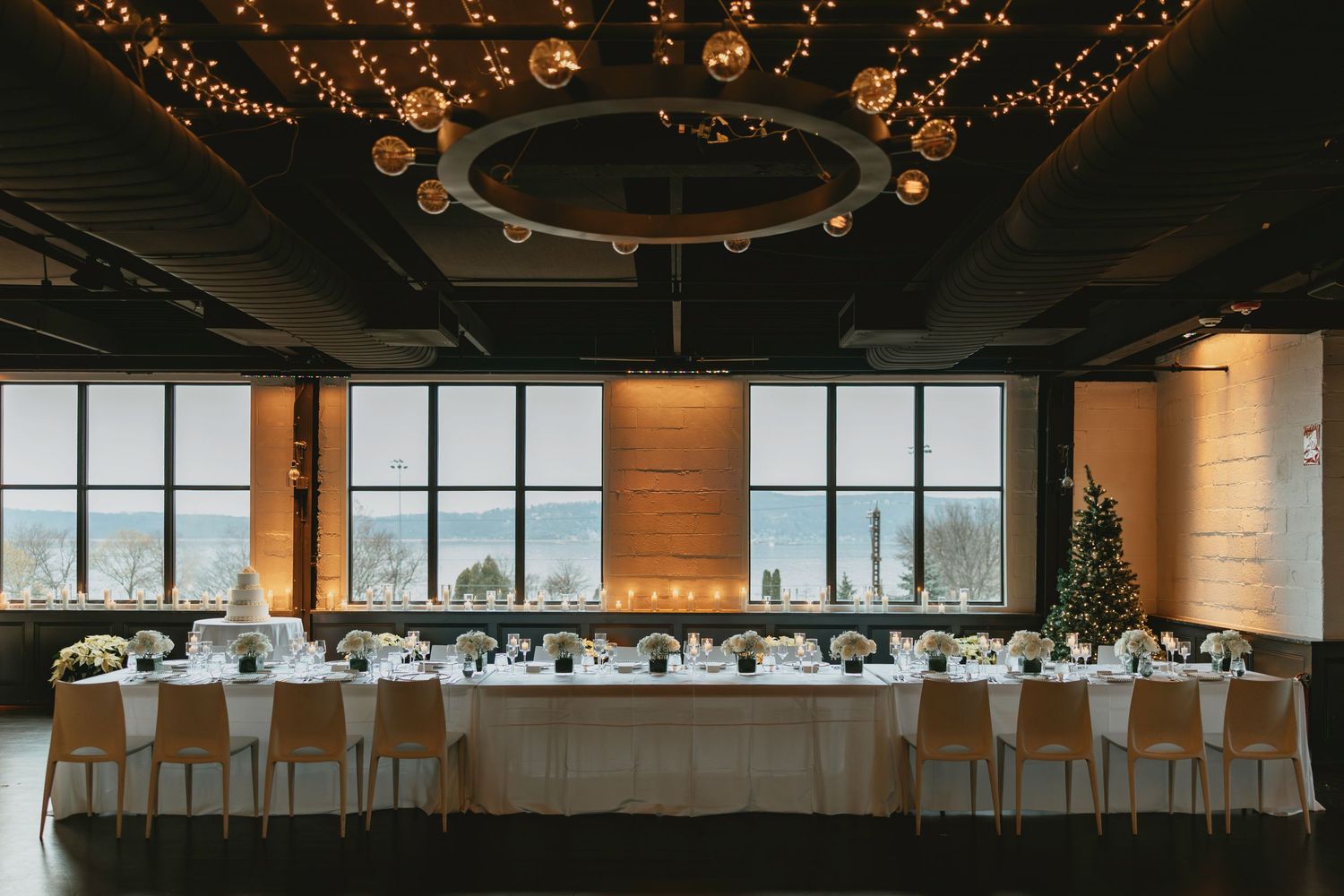 Elegant winter wedding reception with long dining table, string lights, globe pendants, and panoramic windows overlooking a snowy landscape.