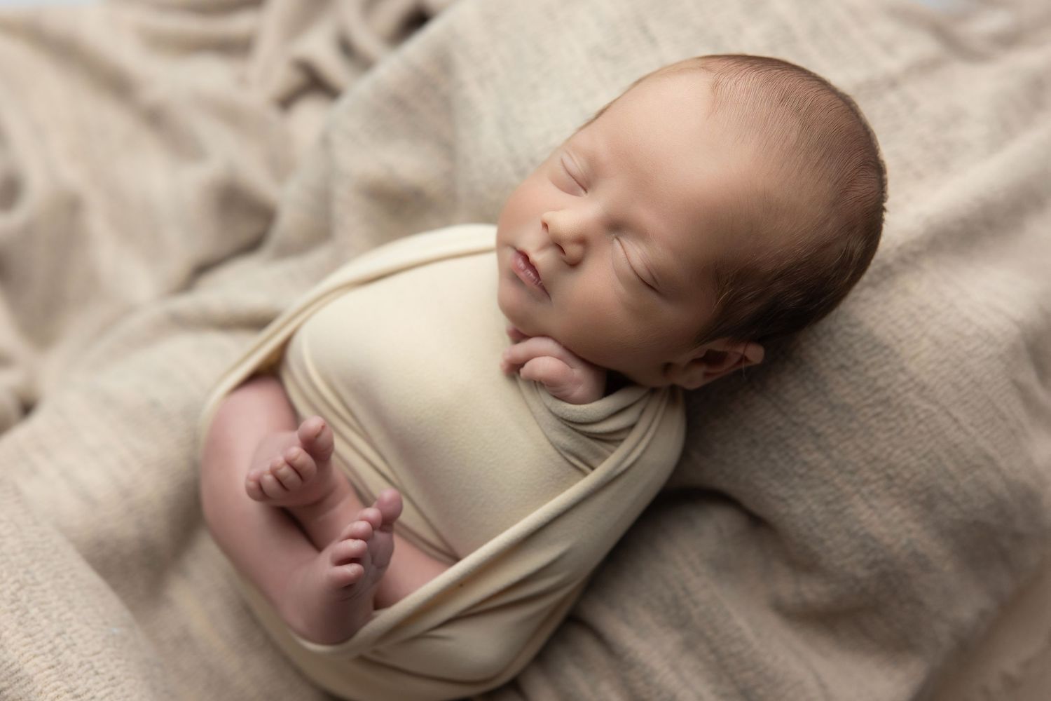 Peaceful newborn baby swaddled in cream wrap sleeping on soft beige blanket during professional newborn photography session.