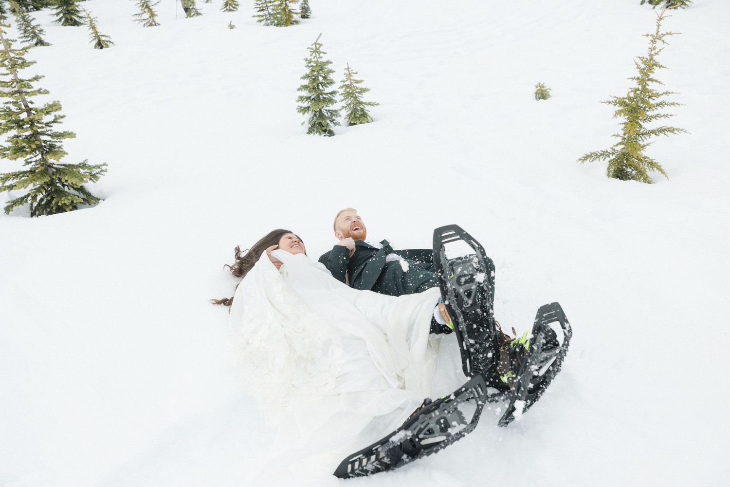 Bride and groom laughing in deep snow during a winter wedding, surrounded by snow-covered evergreen trees.