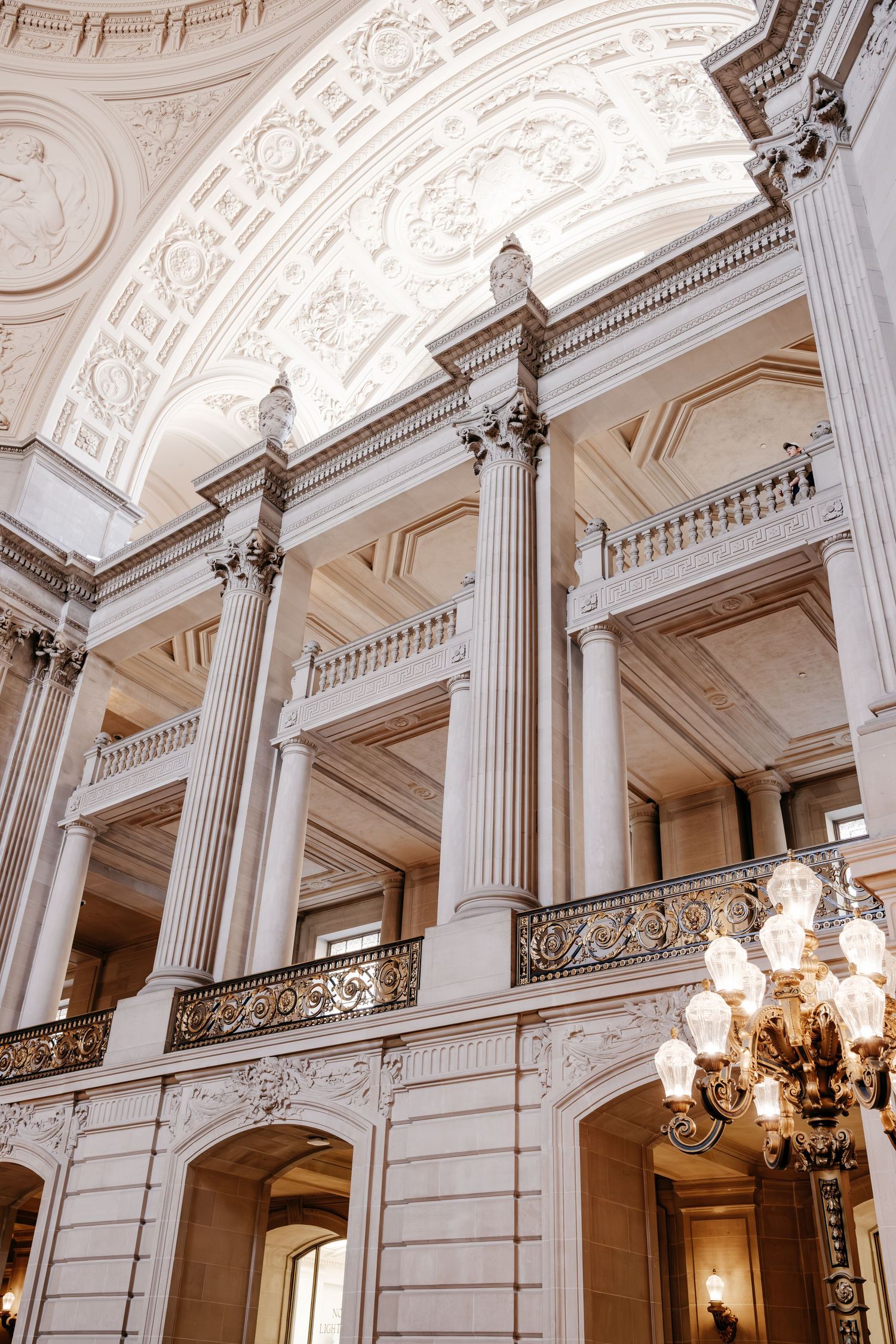 Grand neoclassical interior with ornate columns, arched ceilings, gold balustrades, and elegant chandelier in a historic building.