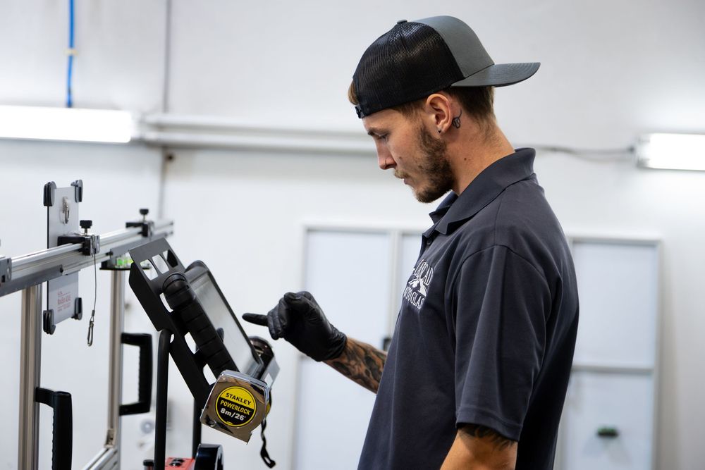 Tattooed mechanic in black polo and cap uses a tape measure while working on metal equipment in a workshop.