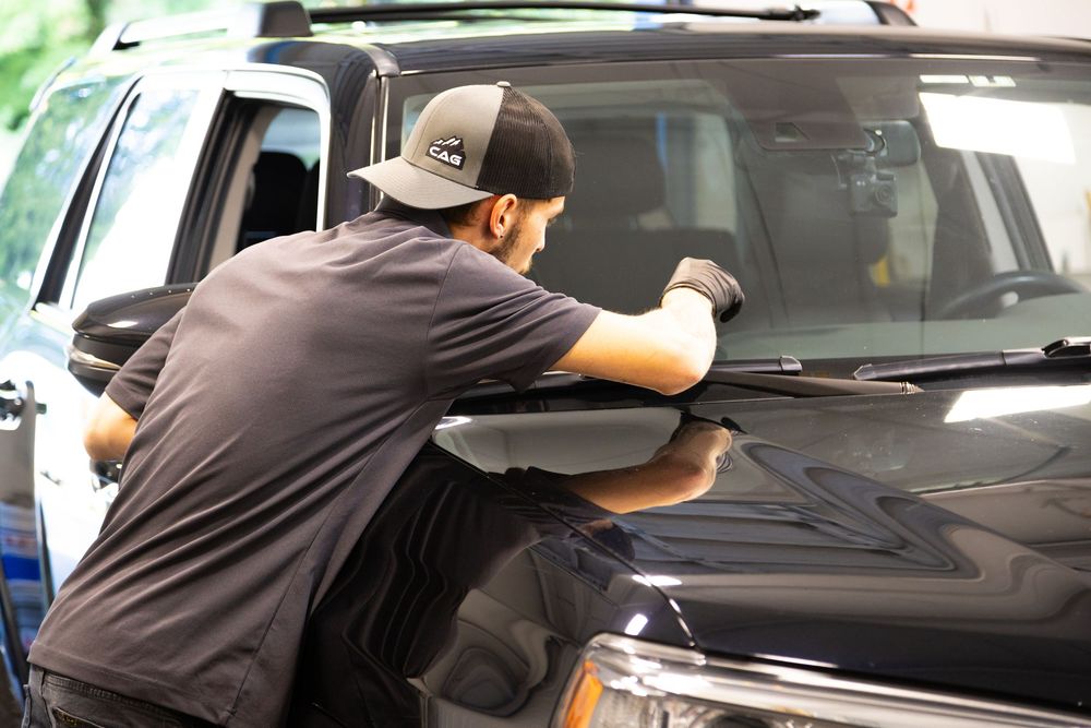 A technician in a cap applies window tint film to a black SUV windshield in an auto detailing shop.