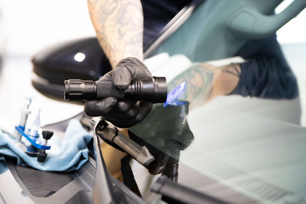 Technician with tattooed arms using UV light tool to repair car windshield crack in auto glass service.