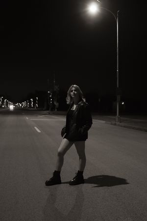 Blonde woman in black jacket and boots standing on empty road under street light at night.