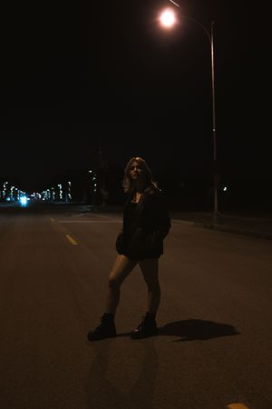 Young woman in black jacket and boots standing on a dark road under a bright street lamp at night.