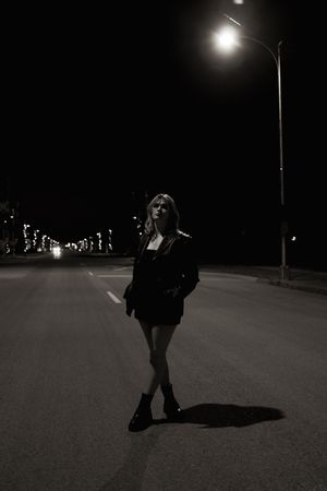 Blonde woman in black mini dress and boots posing on empty road under street lamp at night.