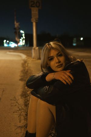 Blonde woman in leather jacket sitting on a curb at night near a speed limit sign, hugging herself.
