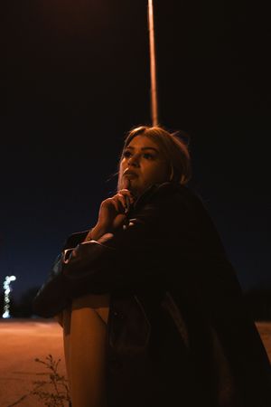 Young woman sitting outdoors at night under a streetlight, wearing a dark jacket, looking contemplative.