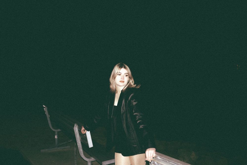 Blonde woman in black leather jacket smiling outdoors at night holding a white object.