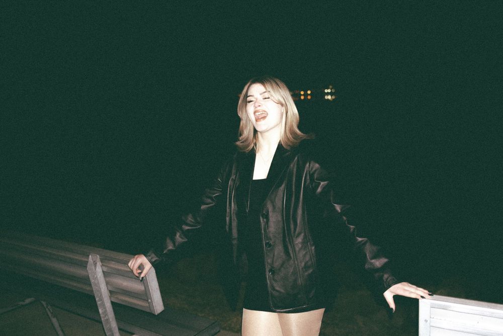 Young woman smiling in a black leather jacket and shorts posing outdoors at night.