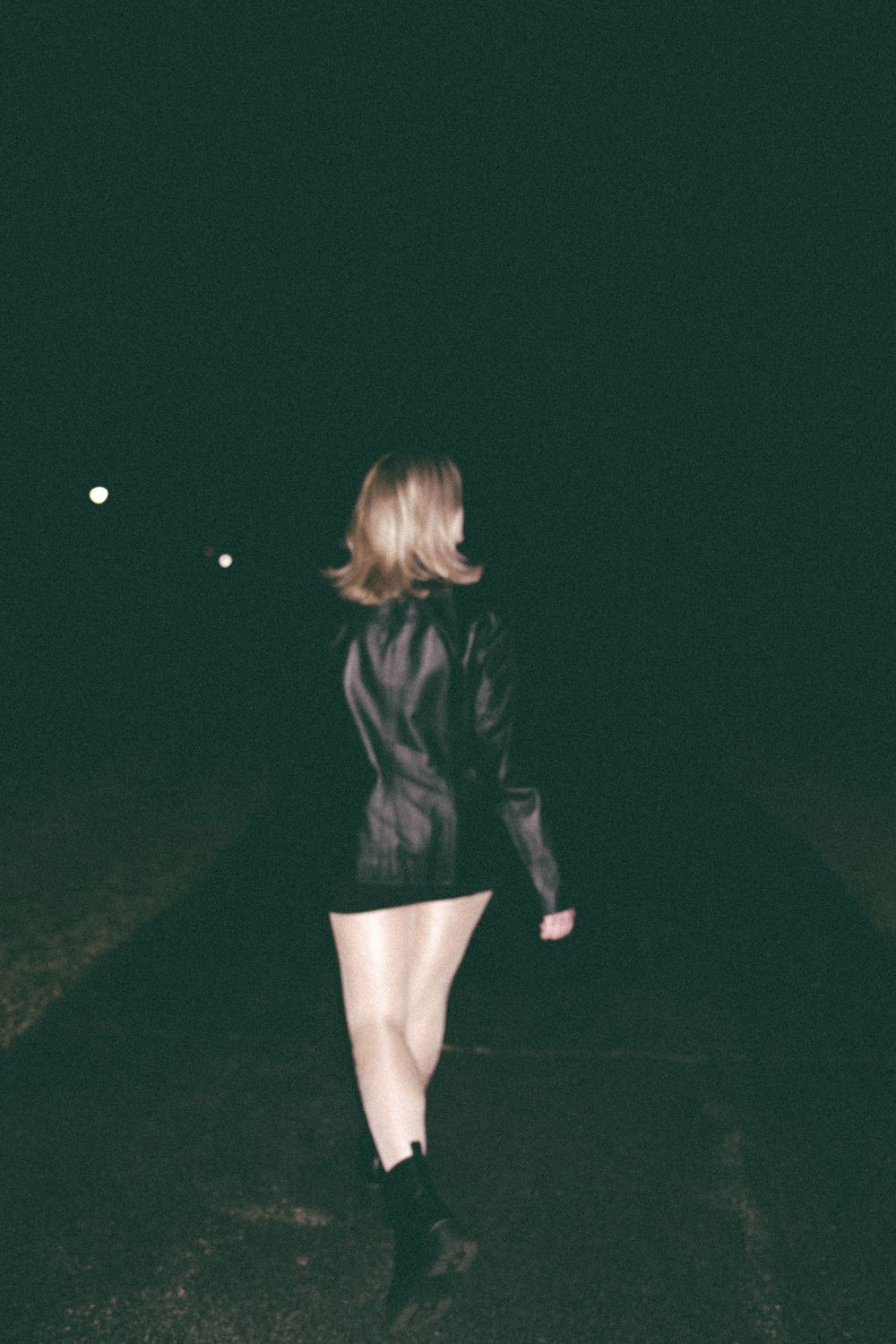 Blonde woman in black leather jacket and mini skirt walking alone on dark road at night.