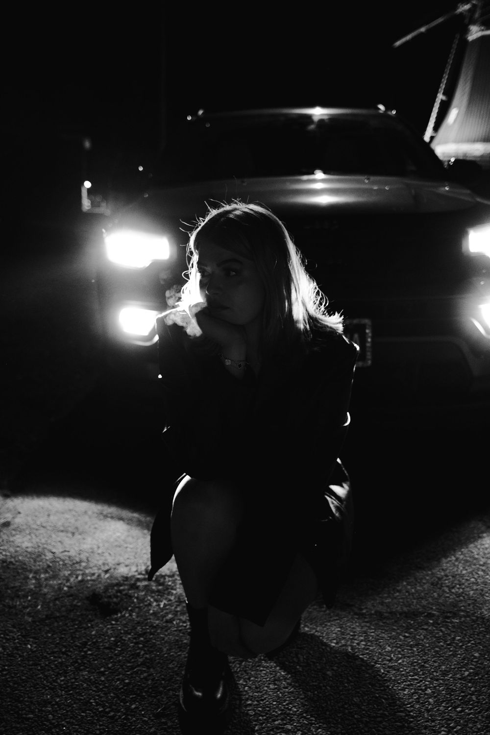 Black and white photo of a blonde woman crouching in front of car headlights at night.
