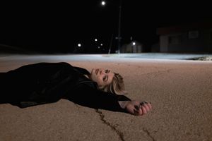 Blonde woman in black jacket lying on empty street pavement under night sky.