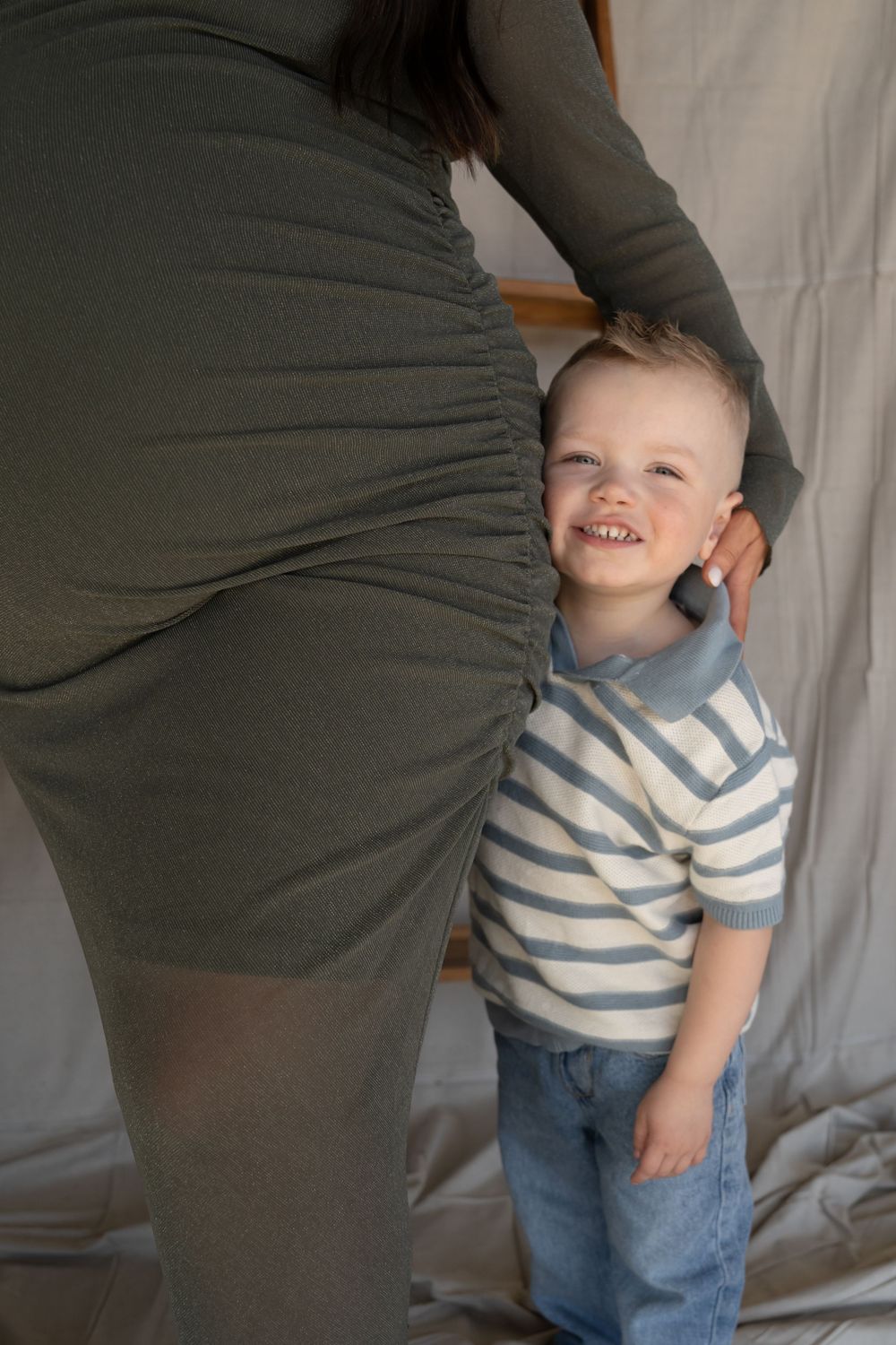 A smiling toddler boy in striped shirt and jeans stands beside a pregnant woman in an olive green dress.