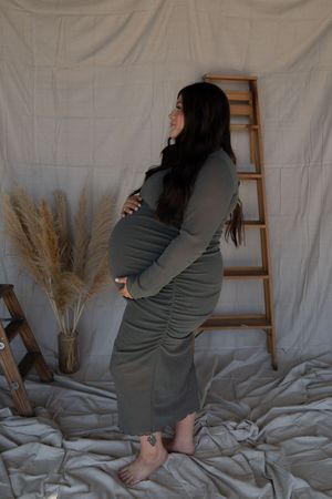 A pregnant woman in an olive ruched dress poses sideways, cradling her bump near pampas grass decor.