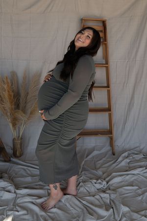 Smiling pregnant woman in olive ruched dress poses barefoot beside wooden ladder in boho maternity shoot.