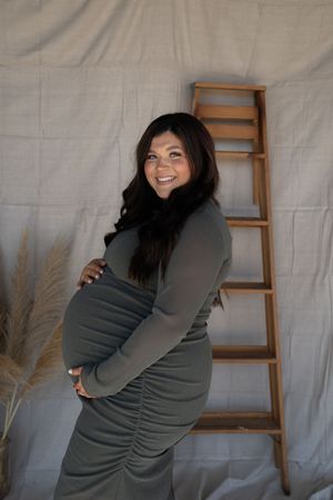 Smiling pregnant woman in olive green ruched maternity dress posing beside wooden ladder with fabric backdrop.