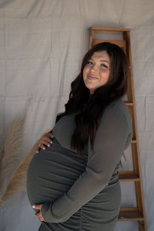 A smiling pregnant woman in a fitted olive green dress cradles her baby bump beside a wooden ladder.