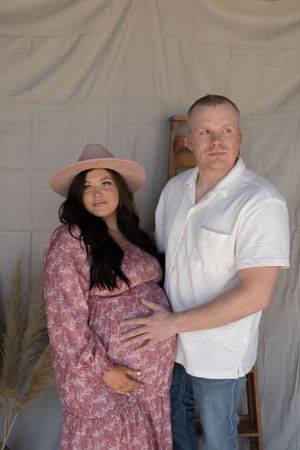 A pregnant couple poses together in a studio, the woman in a floral dress and pink hat, man in white shirt.
