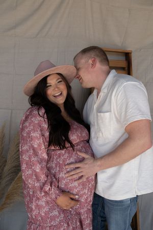 Smiling pregnant woman in pink floral dress and hat with partner touching her baby bump during maternity photo session.