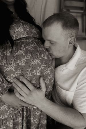 Black and white photo of man tenderly kissing pregnant woman's baby bump in floral maternity dress.