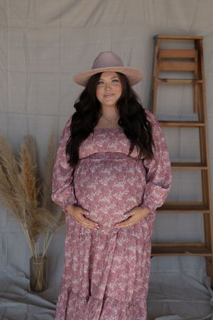 Pregnant woman in pink floral maxi dress and wide-brim hat poses beside wooden ladder in boho maternity shoot.