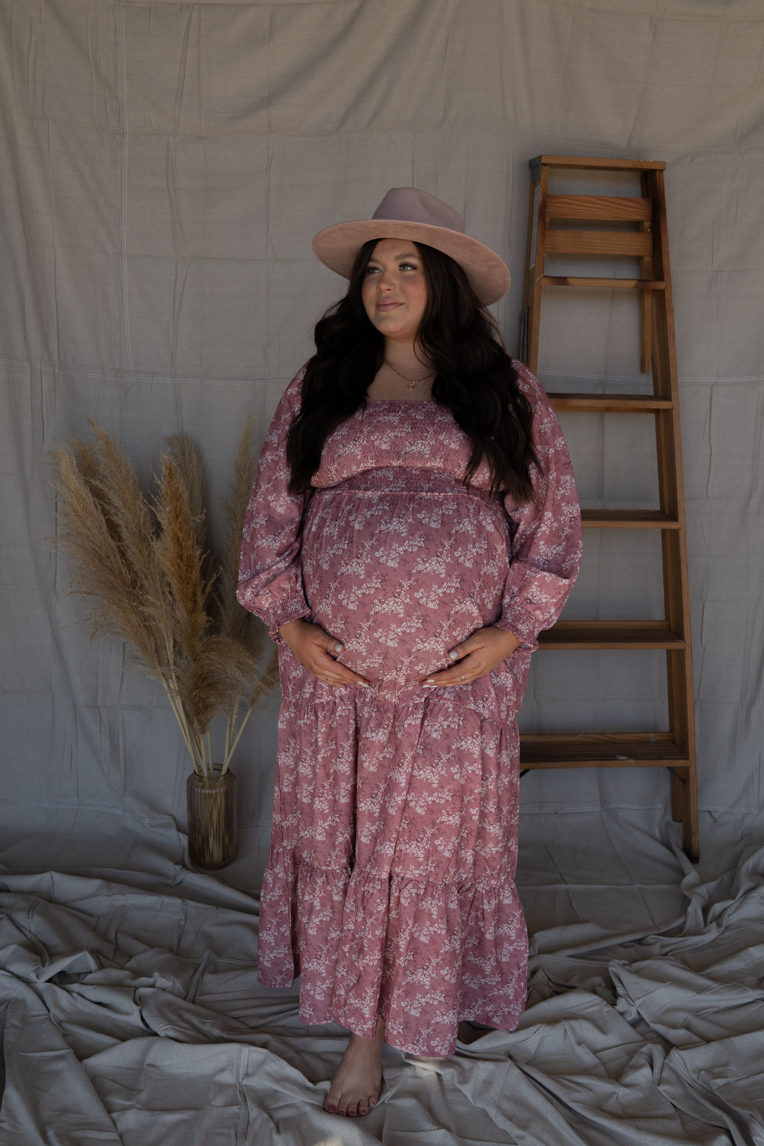 A pregnant woman in a pink floral maxi dress and hat poses gracefully in a neutral-toned studio setting.