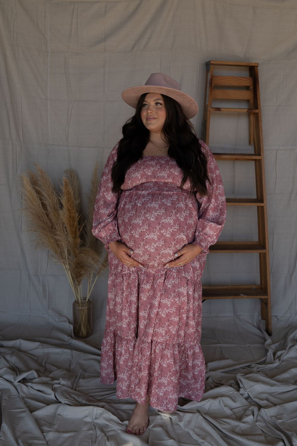 A pregnant woman in a pink floral maxi dress and hat poses gracefully in a neutral-toned studio setting.