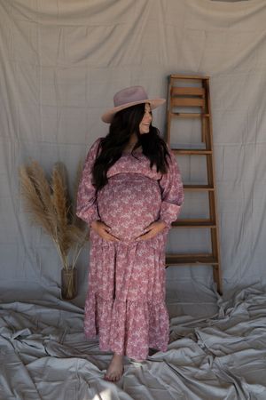 Pregnant woman in floral maxi dress and pink hat poses beside wooden ladder in boho maternity portrait.