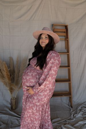 Pregnant woman in pink floral dress and hat posing beside wooden ladder backdrop.