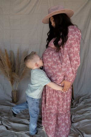 Toddler boy kissing pregnant mother's baby bump during maternity photo shoot, woman in pink floral dress.