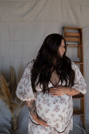 Smiling pregnant woman in floral dress cradles baby bump, long dark hair, tattoos, boho studio setting.