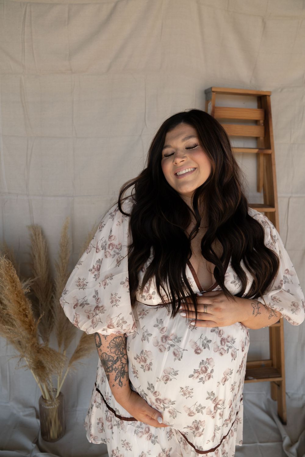 Smiling pregnant woman with long dark hair in floral dress cradling baby bump in soft studio setting.