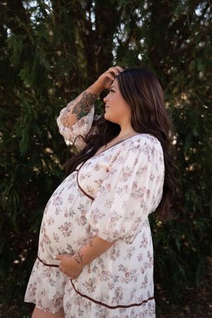 Pregnant woman in white floral dress posing outdoors with hand in hair, baby bump visible, green trees behind.