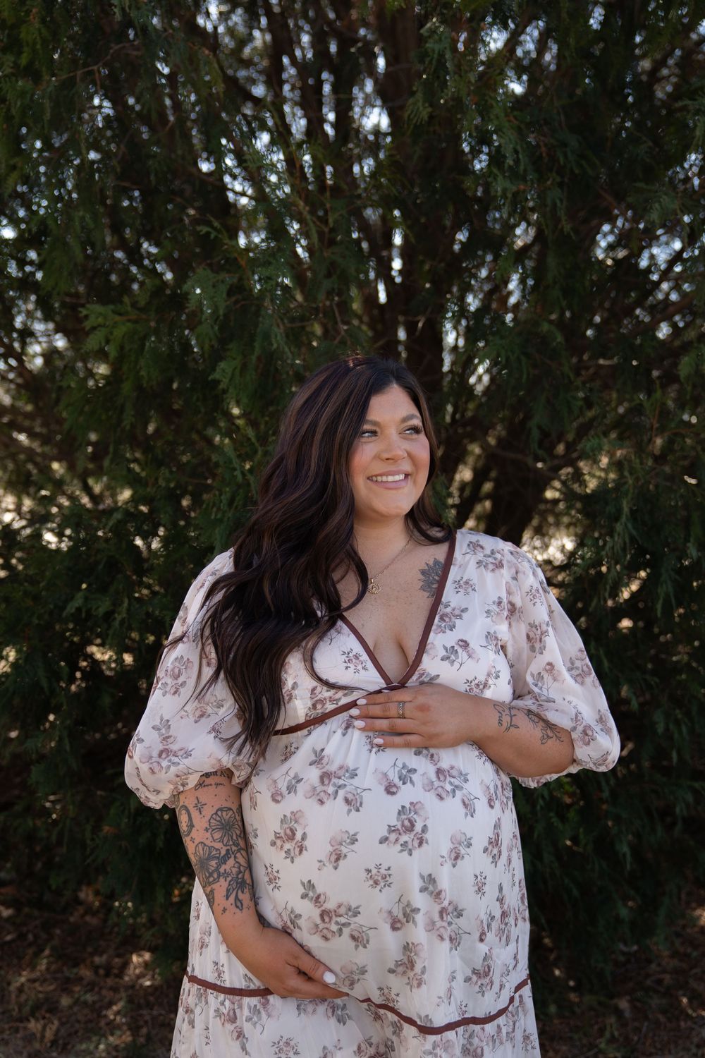 Smiling pregnant woman in floral dress holding baby bump outdoors near evergreen trees.