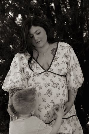 Black and white photo of pregnant woman in floral dress with toddler kissing her baby bump outdoors.
