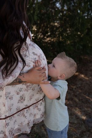 Toddler boy kissing pregnant mothers baby bump outdoors during a maternity photo session.