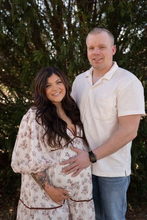 Smiling pregnant couple posing outdoors, woman in floral dress cradling baby bump, man in cream shirt, green trees behind.
