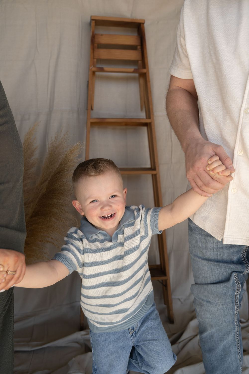 Laughing toddler boy holds hands with parents during a family photo session with rustic ladder backdrop.