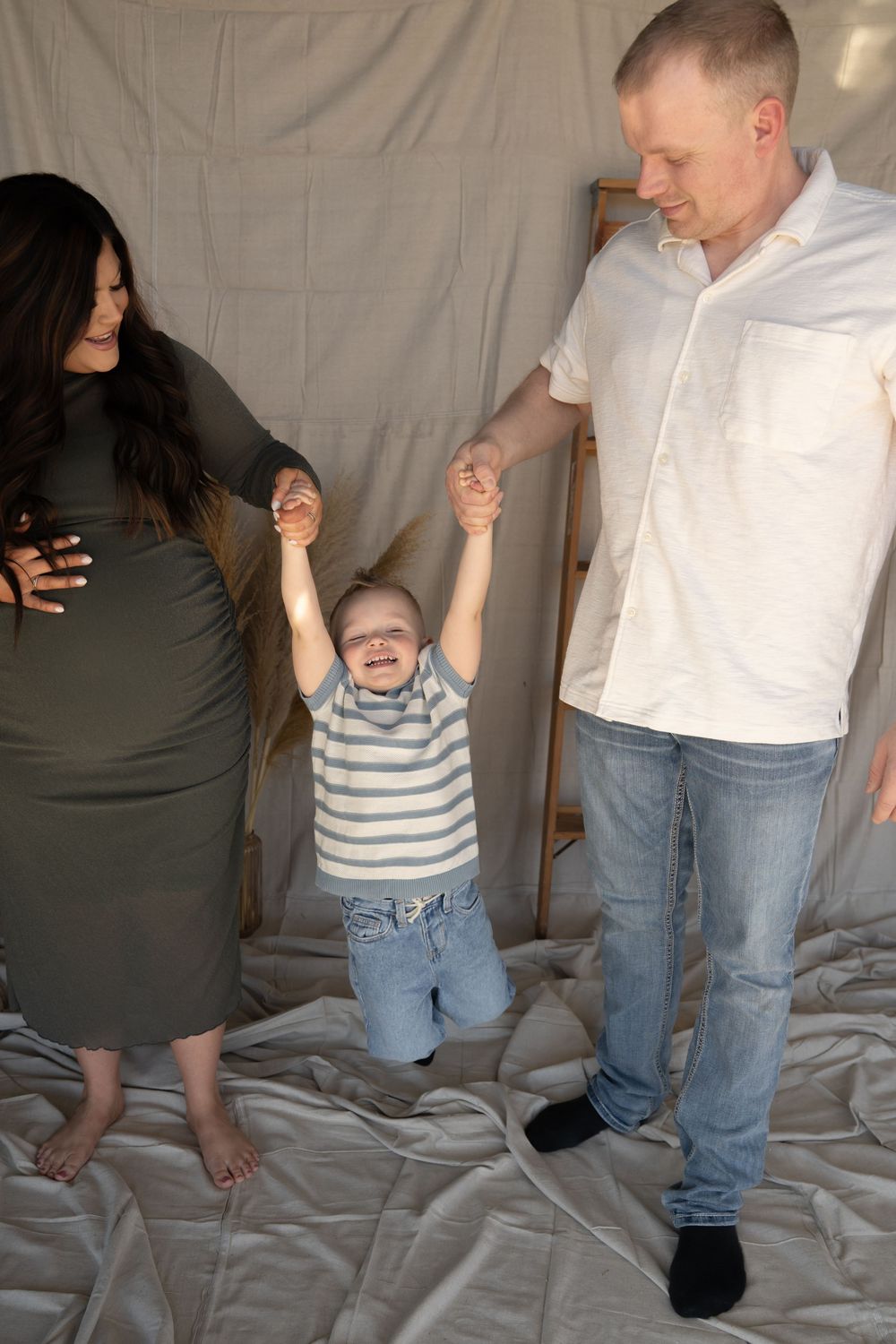 Happy family with pregnant mother, father, and toddler son holding hands in a studio maternity photo session.