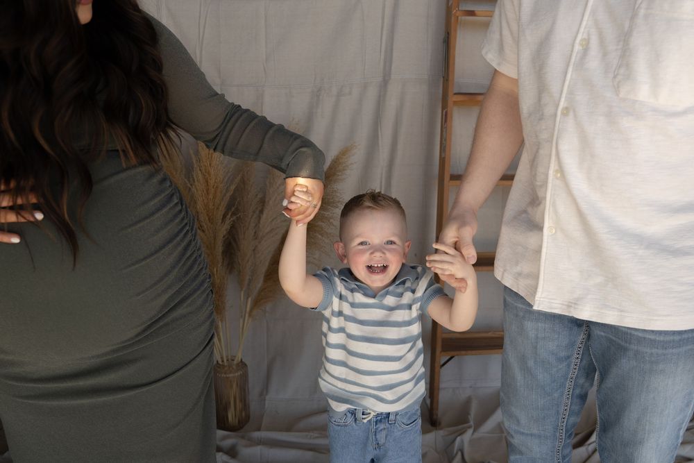 A laughing toddler holds hands with parents during a family photo session against a neutral backdrop.