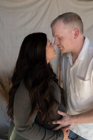 A couple shares an intimate moment, touching foreheads and nearly kissing against a soft white fabric backdrop.