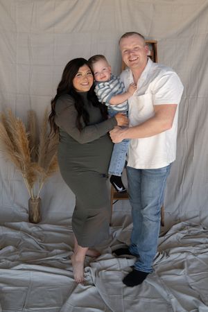 Smiling family maternity photo with pregnant woman in olive dress, man holding baby boy against fabric backdrop.