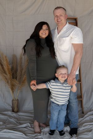 Smiling pregnant family of three with toddler boy in striped shirt posing in neutral studio setting.